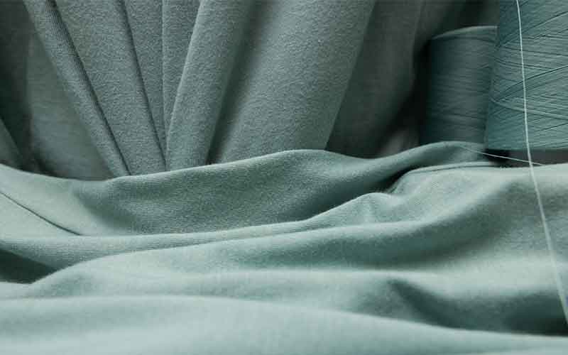 Draped sage green ring-spun cotton jersey fabric with a matching spool of thread, showing the smooth surface of a print-ready blank