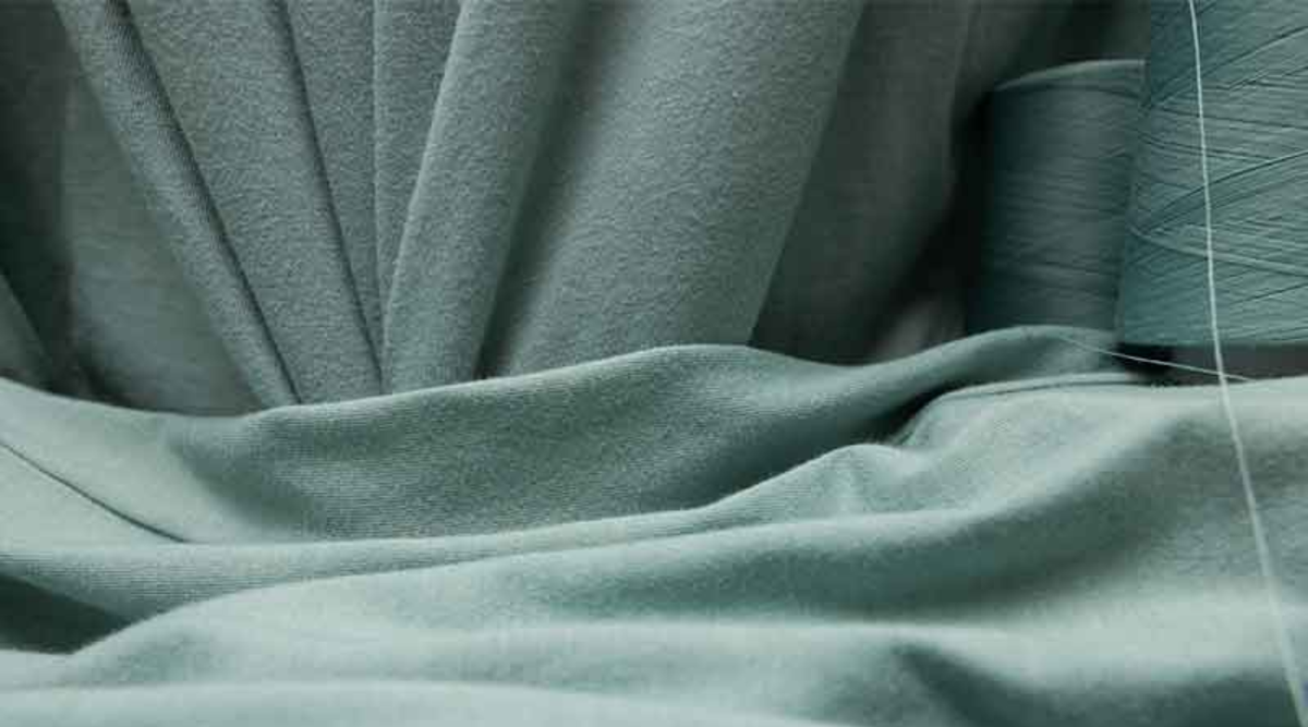 Draped sage green ring-spun cotton jersey fabric with a matching spool of thread, showing the smooth surface of a print-ready blank