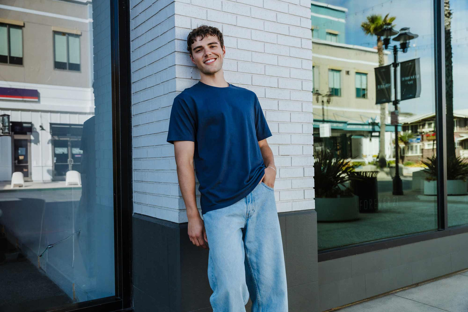 Young man leaning against a white brick wall wearing a plain navy crew-neck blank tee and light-wash jeans
