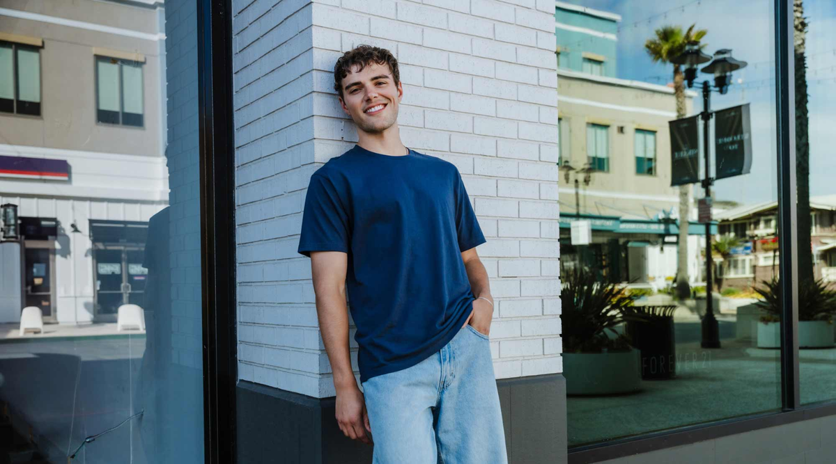 Young man leaning against a white brick wall wearing a plain navy crew-neck blank tee and light-wash jeans