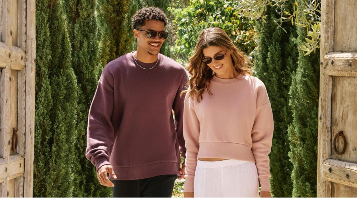 Man in an oversized plum fleece crewneck and woman in a cropped blush pink fleece sweatshirt walking through a sunny garden